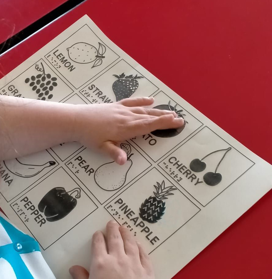 A student explores a tactile table of shapes of fruit with their names written in English in braille and enlarged letters.